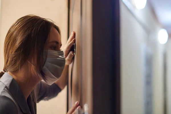 Young woman in medical mask looking through peephole of front door in apartment when somebody rings doorbell. Stay home and self isolation concept. Home quarantine, prevention COVID-19.