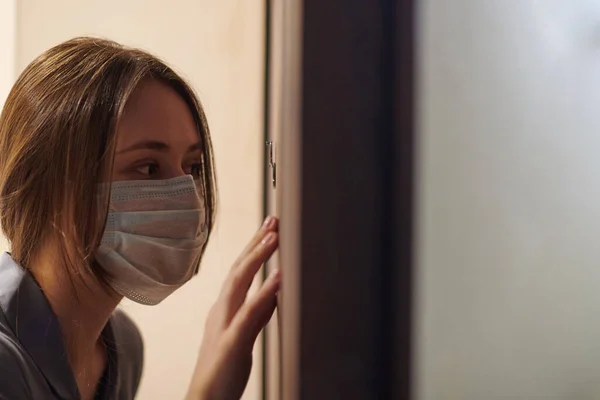 Young woman in medical mask looking through peephole of front door in apartment when somebody rings doorbell. Stay home and self isolation concept. Home quarantine, prevention COVID-19.