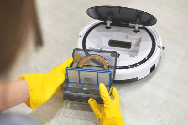 Dust storage box case of robotic vacuum cleaner in gloved female hands. Woman in protective gloves taking out container and filter to clean it from dirt and debris. Vacuum cleaning concept, close up.