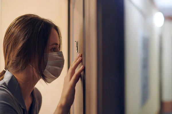 Young woman in medical mask looking through peephole of front door in apartment when somebody rings doorbell. Stay home and self isolation concept. Home quarantine, prevention COVID-19.