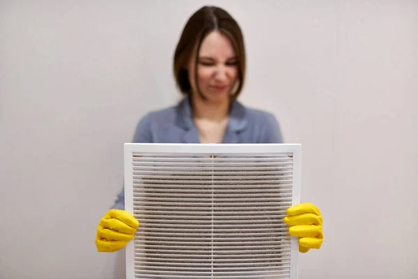 Woman holds ventilation grill with dust filter to clean it. Dirty and dusty white plastic, harmful for health. Cleaning lady with disguised expression, blurred, in protective rubber gloves and uniform