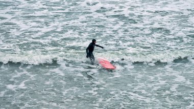 Kaliningrad, Rusya - 01.27.2021 - deniz dalgalarında kırmızı sörf tahtası olan siyah mayo giyen erkek sörfçü. Bulutlu bir gün, mavi dalgalı deniz suları, doğa manzarası. Spor ve açık hava aktivitesi kavramı.