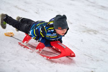 Moskova - 01.12.2021 Renkli ceketli çocuk kırmızı kızağın üstünde kar kaydırağından yuvarlanıyor..