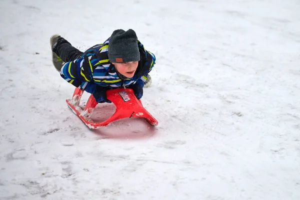 モスクワ 2021 カラフルなジャケットに集中した顔を持つ男の子が赤いそりで雪のスライドを転がしている — ストック写真