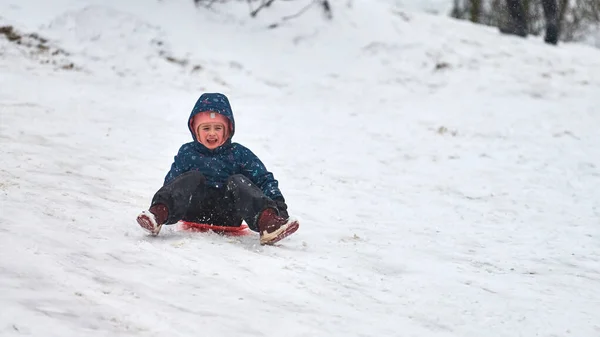 モスクワ 2021 青いダウンジャケットの陽気な女の子が赤いそりで丘を転がっています 冬のスポーツ活動 — ストック写真