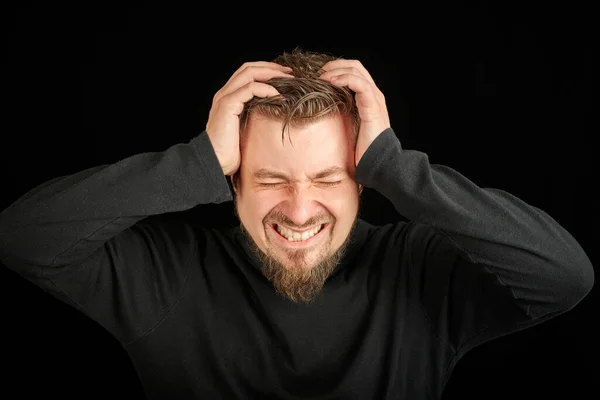 Exhausted bearded man portrait with headache, black background. Overworking, head pain concept. Young guy in black polo neck sweater. Exhausted and tired to point of anger man.