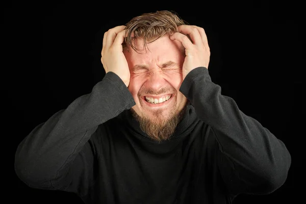 Exhausted bearded man portrait with headache, black background. Overworking, head pain concept. Young guy in black polo neck sweater. Exhausted and tired to point of anger man.