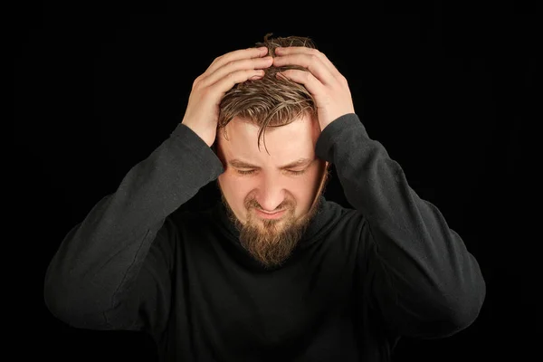 Exhausted bearded man portrait with headache, black background. Overworking, head pain concept. Young guy in black polo neck sweater. Exhausted and tired to point of anger man.