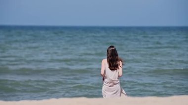Uzun siyah saçlı genç bir kadın açık mavi gökyüzünün altında sakin baltık denizine bakıyor. Manzara sükuneti yumuşak dalgalar ve doğal kıyı güzelliğiyle iletir.
