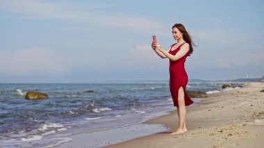 Kırmızı elbiseli genç kadın, Kumlu Baltık Denizi kıyısında çıplak ayakla duruyor, açık mavi gökyüzü ve arka planda dalgalarla selfie çekiyor. Huzurlu kıyı manzaraları sakin suları ve doğal güzellikleri vurgular