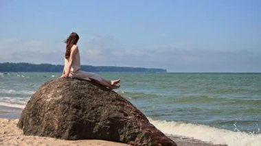 Açık renk elbiseli genç bir kadın, kumlu Baltık Denizi sahilindeki büyük kayalarda oturur, güneşli bir günde, uzak ormanlık kıyı şeridi olan sakin yeşil suları seyreder.