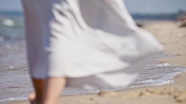 Beyaz elbiseli kadın Baltık Denizi kıyısında ıslak kumda yalınayak yürüyor. Yumuşak doğal ışık altında yumuşak dalgalar sahile vuruyor, sakin ve huzurlu bir atmosfer yaratıyor.