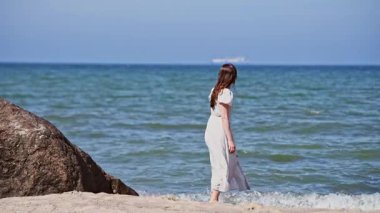 Beyaz elbiseli genç bir kadın açık mavi gökyüzünün altındaki büyük kayaların yakınındaki kumlu Baltık Denizi Sahili 'nde geziniyor. Sakin deniz ve uzak ufuk huzurlu bir kıyı manzarası yaratır.