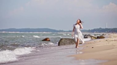 Beyaz elbiseli genç kadın, Baltık Denizi Sahili boyunca yalınayak yürüyor. Yumuşak dalgalar, uzak kıyı şeridi ve kayalarla açık gökyüzü altında ayaklarının yanında dolaşır. Sahne sakin ve huzurlu bir şekilde dışarı çıkar.