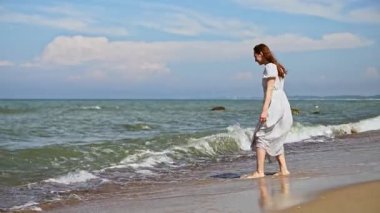 Beyaz elbiseli genç kadın Baltık Denizi kıyısında duruyor, suya dokunmak için öne eğiliyor. Sakin deniz ve yumuşak dalgalar kumlu sahille buluşur açık mavi gökyüzünün altında dağınık bulutlarla