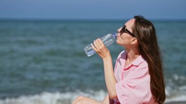 Uzun kahverengi saçlı ve güneş gözlüklü yetişkin bir kadın kumlu sahilde otururken plastik şişeden su içer. Parlak gün ışığı ve mavi okyanus arka planı su ve rahatlamayı vurguluyor