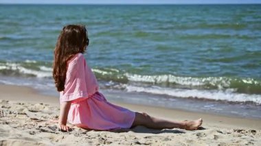 Pembe elbiseli genç bir kadın Baltık Denizi 'nin kumlu sahillerinde oturur ve güneşli bir günde hafifçe dalgalanan suya bakar. Sahne, deniz kenarındaki çevreyi ve sakin dalgaları vurgulayan doğal ışık ile sükuneti yakalar