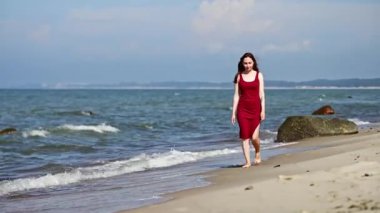 Kırmızı elbiseli genç bir kadın, parçalı bulutlu gökyüzünün altında, Baltık Denizi sahillerinde yalınayak yürüyor. Sakin dalgalar arka planda dağınık kayalarla yavaşça kıyıya vurur.