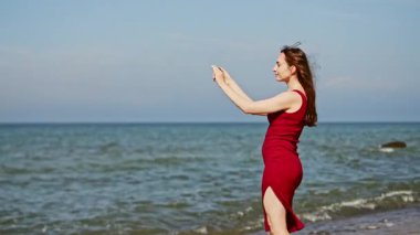 Uzun kahverengi saçlı genç kadın Baltık Denizi sahilinde duruyor, akıllı telefonuyla selfie çekiyor. Sahne sakin sular, berrak gökyüzü ve huzurlu deniz kenarında doğal aydınlatma içeriyor.