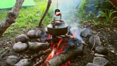 Siyah metal çaydanlık, yeşil orman çevresinde kayalar ve dumanla çevrili kamp ateşinin üzerine asılmış. Sıcak ateş ısıtıcısı, kırsal açık havada atmosfer yaratıyor.