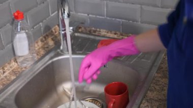 Pembe lastik eldiven giyen eller, mutfak lavabosunda akan suyun altında camları tamamen yıkıyor. Etrafı gri fayanslarla çevrili. Arka planda sabun şişesi ve temiz tabaklar görünüyor.