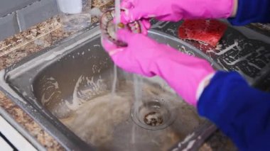 Parlak pembe eldivenler giyen temiz paslanmaz çelik mutfak lavabosu akan suyun altında granit tezgah ve yumuşak ışıklandırmada serpiştirilmiş döşeme