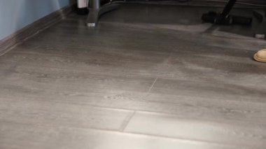 Evin içindeki karanlık ahşap zeminde çekilen süpürme hareketi. Yumuşak ışıklandırma temiz yüzeye vurgu yapar, kişinin rahat terlikleri görünür.