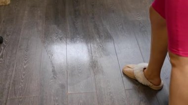 Parlak pembe tayt giyen kadın pencerenin yanındaki koyu ahşap döşemeleri temizlemek için düz paspas kullanıyor. Doğal gün ışığı, yukarıdan gelen ıslak ve kuru zemin kontrastını vurgular