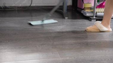 Açık mavi paspas başı aktif olarak parlak koyu ahşap zemini temizliyor. Arka planda mobilya bacakları ve temizlik malzemeleri, temizlik ve bakım vurgulanıyor