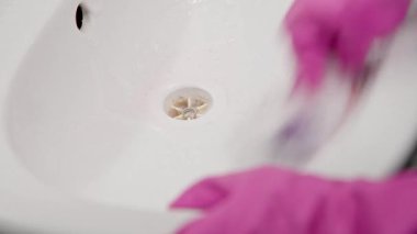 Elleri pembe eldivenlerle yakın plan beyaz lavabo lekesini görünür köpük köpükleriyle ve hafif lekelerle temizleyin. Yumuşak ışıklandırma, ev ortamındaki hijyenik çabaları vurguluyor