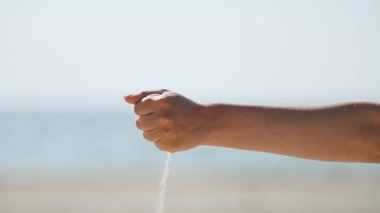 Bronzlaşmış yetişkin kolu yatay olarak tutularak ince düşen kum tanecikleri serbest bırakarak bulanık deniz ve gökyüzü zemininde yavaşça aşağı doğru süzülüyor.