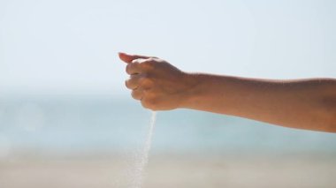 Orta tenli, güneşli yetişkin eli ince kum taneciklerini yavaşça aşağı döküyor, dikey olarak yumuşak mavi gökyüzü ve arka planda bulanık deniz ufku.