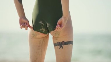 Zeytin yeşili mayo giymiş çıplak bacaklı kadın güneşli sahilde kumu temizliyor. Üst baldırında yay olan dantel jartiyer dövmesine ve alt bacaklarında kumlu dokulara odaklan.