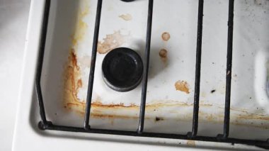 Paslı lekeler ve dağınık yiyecek kalıntıları siyah metal ızgaraların altındaki beyaz metal ocağı kaplıyor. Yakın çekim perspektifi kiri ve lekeli hob yüzeyindeki kiri eşit olmayan ışıklandırmayla vurgular