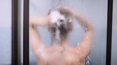 Blurred image of woman washing hair behind foggy, water-spotted glass in tiled shower stall. Soft light and muted colors create intimate, calming atmosphere