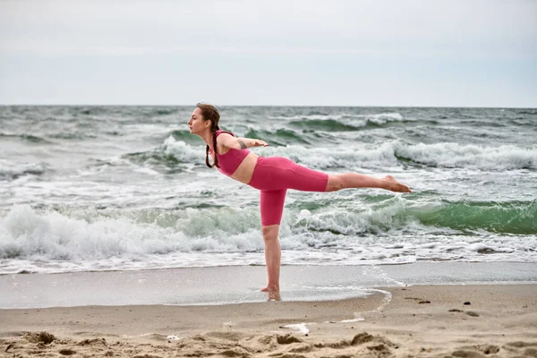 Pembe elbiseli, baltık deniz sahilinde yoga pozu veren genç bir kadın. Dalgaların çarpmasıyla bulutlu gökyüzü dinamik bir zemin oluşturur. Sakin odaklanma, doğal enerjiyle çelişir.