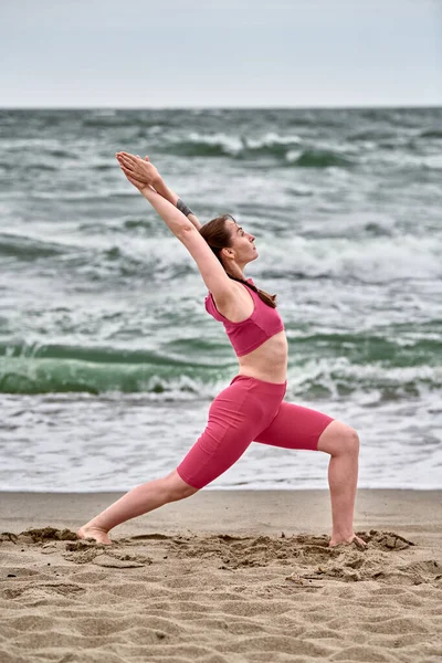 Baltık Sahili 'nde canlı pembe giysiler içinde yoga yapan genç bir kadın. Dinamik savaşçı durgun deniz dalgaları ve bulutlu gökyüzü arasında huzur dolu bir ortam yaratır.