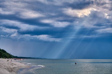 Baltık Denizi kıyısında fırtına bulutlarını aşan güneş ışınlarıyla dramatik bir gökyüzü. Kum kıyıları suyu özetlerken, uzak ağaçlar yoğun atmosfere zıtlık ekler.