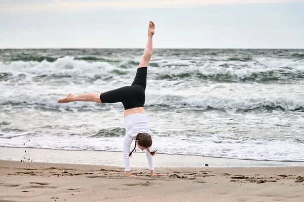 Genç bayan, kumlu Baltık Sahili 'nde takla atıyor. Bulutlu gökyüzü ve dalgalar, denge ve özgürlüğü vurgulayarak sakin deniz kenarı ortamını tanımlar. Yumuşak, sessiz tonlar doğal güzelliği vurgular