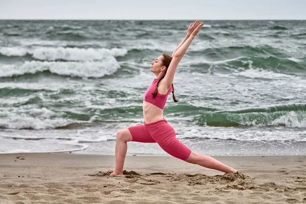 Baltık Sahili 'nde canlı pembe giysiler içinde yoga yapan genç bir kadın. Dinamik savaşçı durgun deniz dalgaları ve bulutlu gökyüzü arasında huzur dolu bir ortam yaratır.
