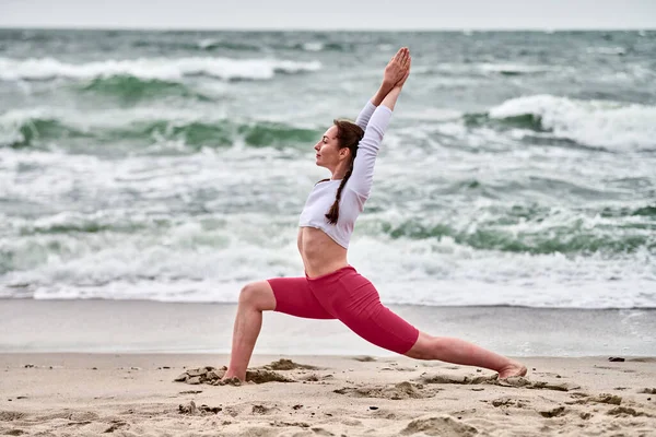 Kumlu Baltık sahilinde yoga yapan genç bir kadın. Bulutlu gökyüzü sakin bir atmosfer yaratır. Pembe şort giyiyor, beyaz bluz giyiyor, savaşçı pozu veriyor.