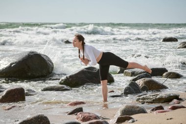 Genç bayan kayalık baltık deniz kıyısında yoga pozu veriyor. Onun odaklanmış ifadesi çalkantılı dalgalarla çelişiyor. Bulutlu gökyüzü dingin atmosfer ekler