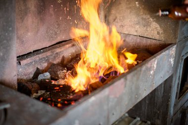 Parlak turuncu alev, koyu metalle çevrili demirhaneden sıçrar. Kıvılcımlar uçuşuyor, atölye ortamının canlı enerjisini gösteriyor. Yakın çekim yoğunluğu yakalar