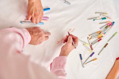 Beyaz kağıt üzerinde renkli kalemlerle çizim yapan iki elin yakın çekimi. Etrafa dağılmış çeşitli işaretler yaratıcı ve sıradan atmosfer yaratır.