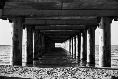 Baltık Denizi rıhtımının altındaki siyah beyaz manzara doğal gün ışığı altında durgun sulara doğru uzanan simetrik beton sütunların tünel benzeri bir perspektif oluşturduğunu gösteriyor.