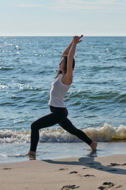 Genç bir kadın gün doğumunda Baltık Denizi kıyısında çıplak ayakla yoga yapıyor. Sakin su ve huzurlu farkındalık, sakin bir açık hava ortamı yaratır.