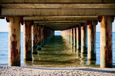 Sakin Baltık Denizi sularında duran paslı sütunlarla yıpranmış iskele manzarası. Yumuşak altın ışık, kumlu kıyılarla çelişen mavi-yeşil renkleri güçlendirir.
