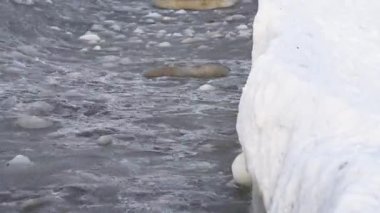 Yoğun kar yığınının yanında soğuk gri su üzerinde yüzen desenli buzlu buz tabakası, düşük açı görüntüsü buzlu desenleri ve donmuş kıyı ortamını vurguluyor.