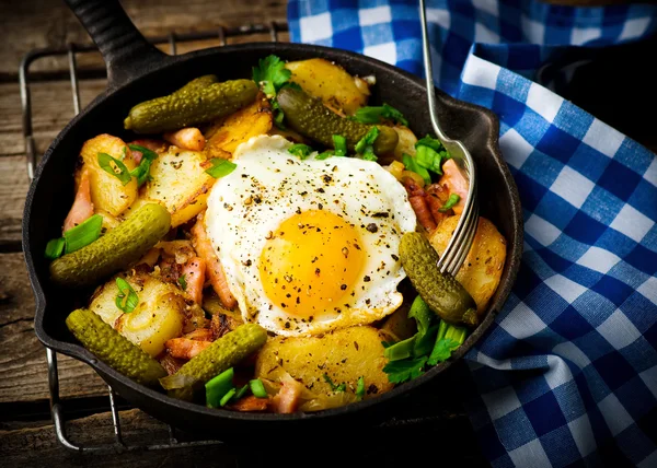 Country breakfast from potatoes, with bacon and fried eggs in a pig-iron frying pan. — Stock Photo, Image