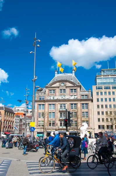 АМСТЕРДАМ-АПРЕЛЬ 30: Неопределенные люди переходят улицу на велосипедах на площади Дам, флагманский магазин De Bijenkorf виден на заднем плане 30 апреля 2015 года в Амстердаме, Нидерланды . — стоковое фото